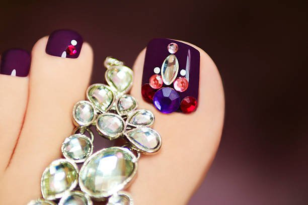 Elegant pedicure with rhinestones on purple nail varnish female foot on a dark background.